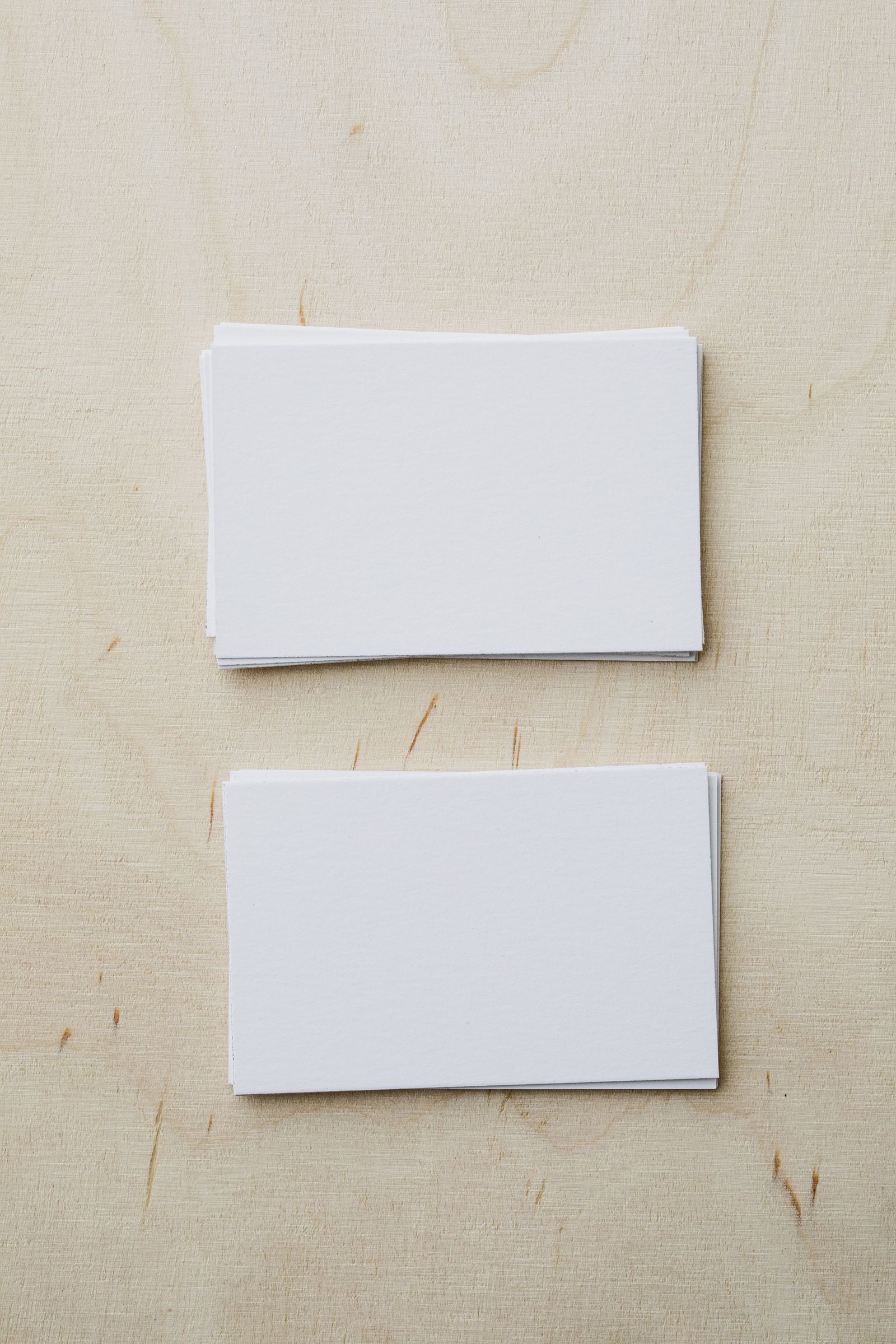 Two stacks of blank white business cards on a pale plywood surface, viewed from above.