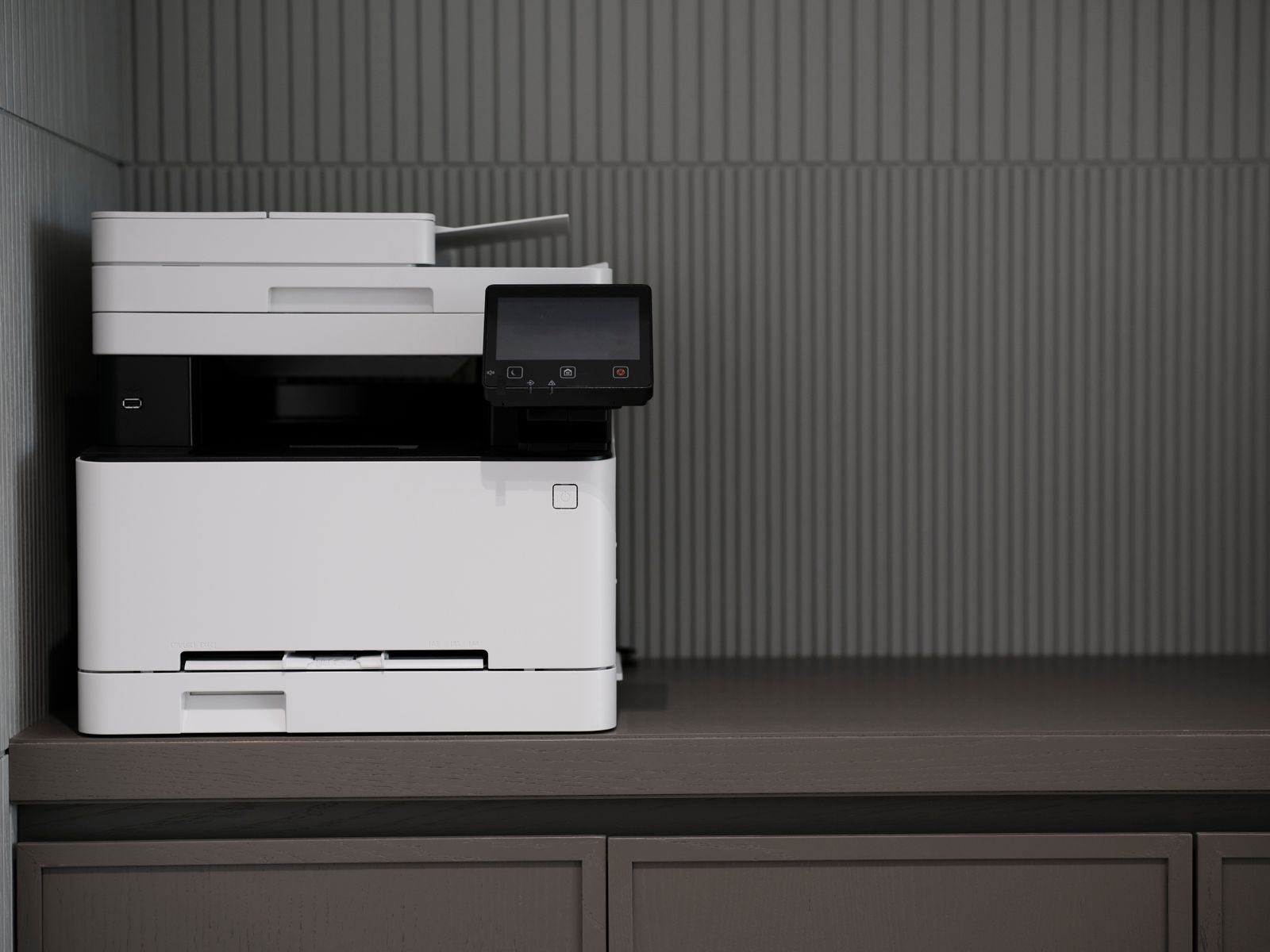A white-and-black multifunction printer with a small touchscreen, standing on a grey-brown cabinet top against a ribbed grey wall.